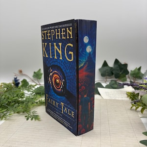Könnte beinhalten: Ein Hardcover-Buch mit dem Titel "Fairy Tale" von Stephen King. Das Cover zeigt ein blau-goldenes Design mit einem zentralen augenartigen Bild. Der Buchrücken zeigt eine Nachtszene mit einer Burg und zwei Monden.