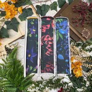 May include: Three book covers with floral designs. The covers feature a dark background with vibrant flowers in purple, red, and blue. The covers have a silver metal frame. The books are surrounded by greenery, flowers, and open books.