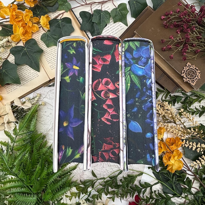May include: Three book covers with floral designs. The covers feature a dark background with vibrant flowers in purple, red, and blue. The covers have a silver metal frame. The books are surrounded by greenery, flowers, and open books.