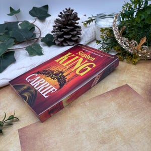 Könnte beinhalten: Ein Hardcover-Buch mit dem Titel "Carrie" von Stephen King. Das Buch hat einen rot-orangen Einband mit dem Titel in Weiß und Gelb. Das Buch liegt auf einer Holzoberfläche mit dekorativen Elementen.
