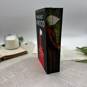 May include: A hardcover edition of Margaret Atwood's "The Handmaid's Tale" is displayed. The book's cover features a woman in a red dress and white bonnet. The title and author's name are visible. The book is set against a backdrop of printed paper and a candle.