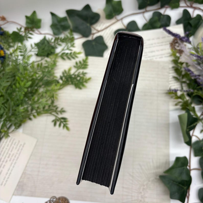 May include: A hardcover book with a black cover and black-edged pages. The spine is visible, showing the book's thickness. The book is surrounded by greenery and floral elements, suggesting a decorative or aesthetic presentation.