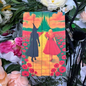 Puede incluir: Panel de madera con una colorida ilustración de una escena de El Mago de Oz. La obra representa a dos figuras caminando por un camino de ladrillos amarillos hacia un castillo verde, con amapolas rojas.