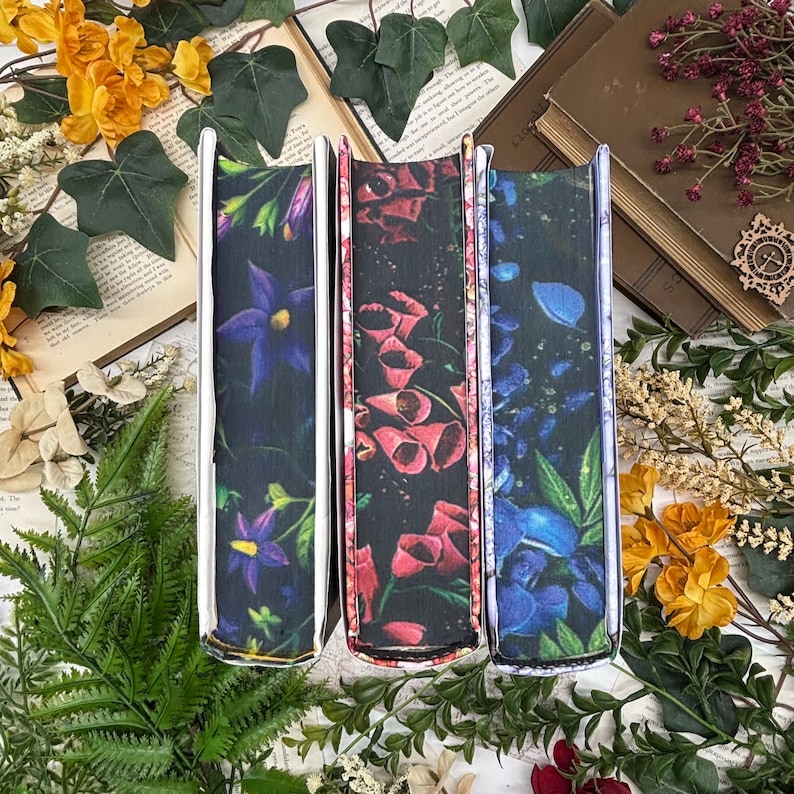 May include: Three book covers with floral designs. The spines feature dark backgrounds with vibrant flowers in purple, red, and blue. The covers are surrounded by greenery, yellow flowers, and open books.