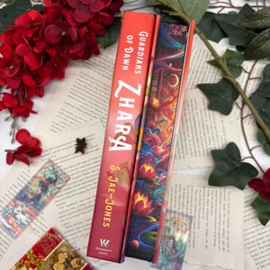 May include: A red hardcover book titled "Guardians of Dawn Zharas" by S. Jae-Jones. The book is part of the Wednesday Books series. The book cover features a colorful illustration of a fantasy scene with a dragon and a woman.
