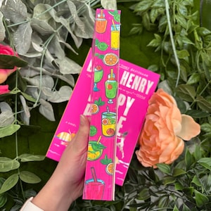 May include: A pink book with the title "Henry Happy" is displayed. The spine features colorful illustrations of various beverages, including orange slices, limes, and straws. The book is surrounded by greenery and a peach-colored flower.