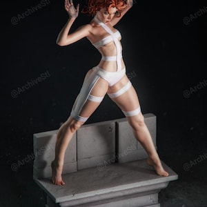 May include: A white figurine of a woman with red hair, wearing a white outfit with straps. The figure is posed with arms raised, standing on a grey stone platform against a dark background. The figurine is a detailed sculpture.