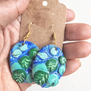 May include: A pair of blue and green painted clay earrings with gold hooks. The earrings feature a swirling pattern with green leaves and white dots.