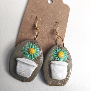 May include: A pair of ceramic earrings shaped like a flower pot with a yellow flower on top. The earrings are brown with white accents and have gold hooks.