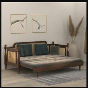 May include: A dark wood daybed featuring a woven cane back and side panels, a tufted gray mattress, and two dark teal square throw pillows. The frame includes turned wooden posts at the corners and short, tapered legs supporting the base structure.