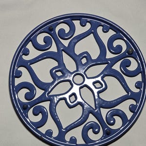 May include: A circular blue trivet featuring an intricate openwork design of symmetrical swirls and heart-shaped patterns. The metal piece has a glossy finish and includes small, dark protective feet positioned along the outer rim for stability.