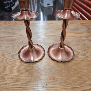 May include: Two copper-colored candle holders with a twisted stem design. Each holder has a scalloped base and a flared top. The candle holders are displayed on a wooden surface.
