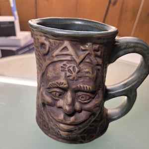 May include: A brown ceramic mug featuring a sculpted face with a wide, smiling expression. The surface has a textured, matte finish with dark blue-green accents along the rim and handle. Various geometric symbols are embossed above the face.