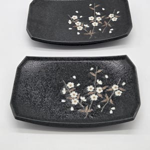 May include: Two rectangular black ceramic plates with curved edges and a textured finish. Each plate features a painted design of white cherry blossoms with brown branches and scattered petals across the dark speckled surface.