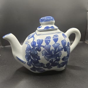 May include: White ceramic teapot with a blue floral pattern. The teapot has a curved spout, a handle, and a lid. The blue floral design covers the body of the teapot, creating a classic and elegant look. The teapot is a decorative item.