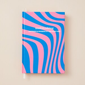 Op de afbeelding: Een hardcover notitieboek met een blauw en roze golvend patroon. De voorkant toont het woord "multitasking" in het wit, met de zin "listening & ignoring at the same time" eronder. Een wit lint als bladwijzer is zichtbaar.