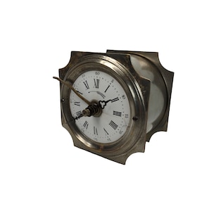May include: A silver-toned clock with Roman numeral markings on a white face. The clock has a decorative design around the edge and a small, decorative wheat stalk above the XII.