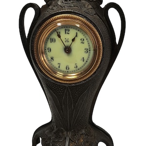 Könnte beinhalten: Eine schwarze gusseiserne Kaminuhr mit einem goldenen Zifferblatt und floralen Akzenten. Die Uhr hat zwei Griffe und einen dekorativen Sockel.