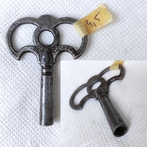 May include: A vintage, dark gray metal clock key with a decorative, openwork design. The key features a circular opening and two curved handles. A small piece of paper with the number "4.5" is taped to the key.