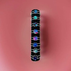 May include: A display of galaxy-themed wire-wrapped bracelets with colourful glass cabochons. The bracelets are arranged vertically on a black stand against a pink background.