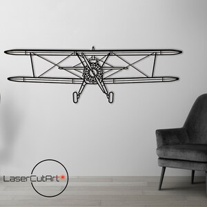 Op de afbeelding: Zwarte metalen wandkunst van een vintage vliegtuig, met een zwarte fauteuil en een zwarte staande lamp met een gouden voet in de achtergrond.