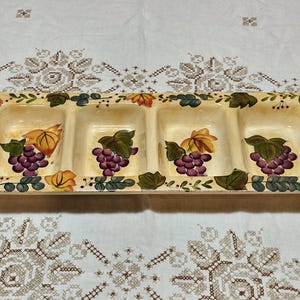 May include: Rectangular serving dish with four compartments. The dish is cream-colored with a painted grape and leaf design in shades of purple, green, and yellow. The edges are decorated with vines and leaves. A decorative piece for serving snacks or appetizers.