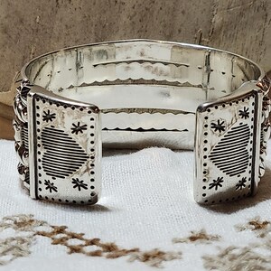 K&ouml;nnte beinhalten: Ein silbernes Armreif-Armband mit offenem Design. Das Armband verf&uuml;gt &uuml;ber dekorative Paneele mit ge&auml;tzten Herz- und Sternmustern. Die Kanten sind mit kleinen, erhabenen Details verziert. Das Armband wird auf einer wei&szlig;en Oberfl&auml;che pr&auml;sentiert.