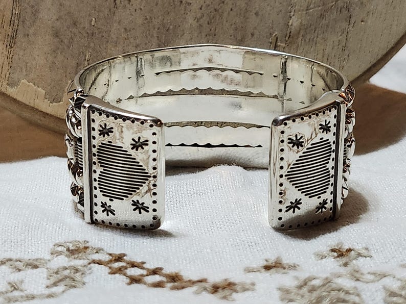 K&ouml;nnte beinhalten: Ein silbernes Armreif-Armband mit offenem Design. Das Armband verf&uuml;gt &uuml;ber dekorative Paneele mit ge&auml;tzten Herz- und Sternmustern. Die Kanten sind mit kleinen, erhabenen Details verziert. Das Armband wird auf einer wei&szlig;en Oberfl&auml;che pr&auml;sentiert.