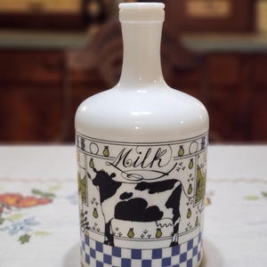 May include: White ceramic milk bottle with a black and white cow illustration. The bottle has a blue and white checkered base and a decorative border with the word "Milk".