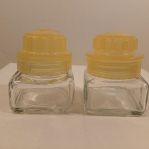 May include: Two clear glass jars with yellow plastic lids. The lids have a ridged design.