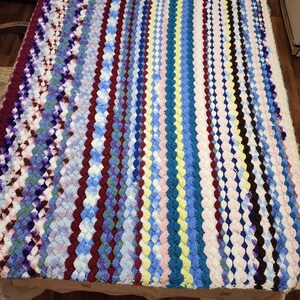 May include: A handmade, colorful crochet blanket. The blanket features vertical stripes in various colors, including blue, purple, red, yellow, and white. The crochet pattern creates a textured, raised design, perfect for a cozy home decor item.