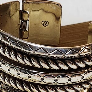 K&ouml;nnte beinhalten: Nahaufnahme eines silberfarbenen, aufklappbaren Armreifs. Das Armband weist aufwendige Designs auf, darunter geometrische Muster und eine seilartige Textur. Der Verschluss ist goldfarben mit einem kleinen Herzsymbol.