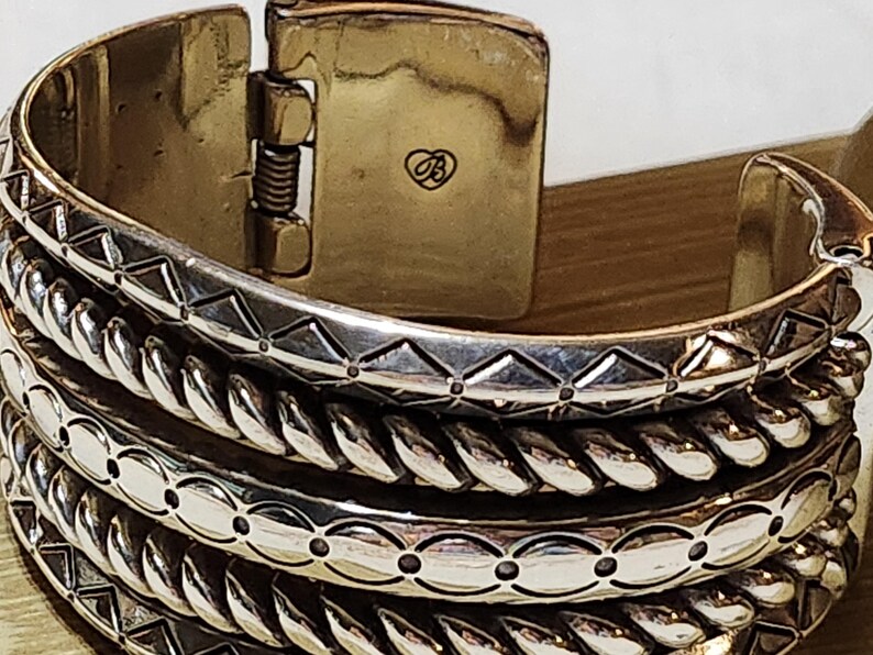 K&ouml;nnte beinhalten: Nahaufnahme eines silberfarbenen, aufklappbaren Armreifs. Das Armband weist aufwendige Designs auf, darunter geometrische Muster und eine seilartige Textur. Der Verschluss ist goldfarben mit einem kleinen Herzsymbol.