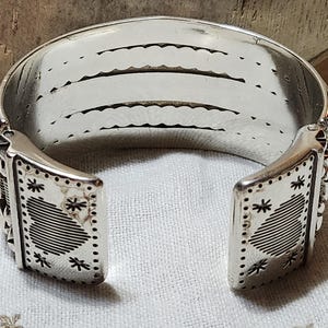 K&ouml;nnte beinhalten: Ein silbernes Armreif-Armband mit offenem Design. Das Armband hat dekorative Stern- und geometrische Muster an den Enden. Der Innenraum hat drei parallele Linien mit einem gewellten Rand. Das Armband ist aus gl&auml;nzendem Metall gefertigt.