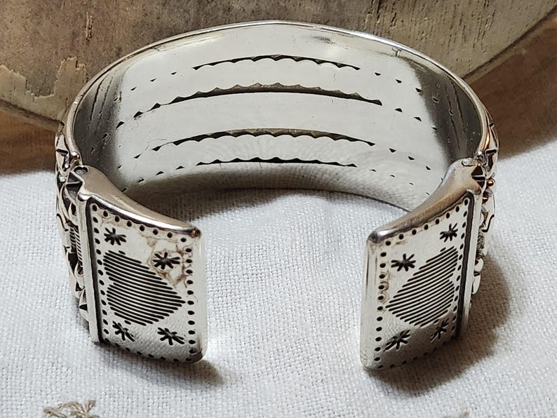 K&ouml;nnte beinhalten: Ein silbernes Armreif-Armband mit offenem Design. Das Armband hat dekorative Stern- und geometrische Muster an den Enden. Der Innenraum hat drei parallele Linien mit einem gewellten Rand. Das Armband ist aus gl&auml;nzendem Metall gefertigt.