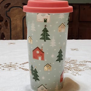 May include: A tall, light blue travel mug with a red lid. The mug is decorated with small houses, Christmas trees, and snowflakes. The mug is designed to keep beverages hot or cold. The mug is made of ceramic and has a silicone lid.