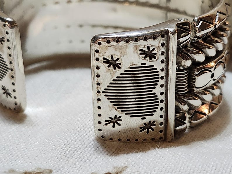 K&ouml;nnte beinhalten: Nahaufnahme eines silbernen Armreifs. Das Armband weist aufwendige Designs auf, darunter Sternmuster und Streifen. Der Verschluss ist mit dekorativen Elementen versehen. Das Gesamtdesign ist aufwendig und detailliert.