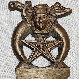 May include: Bronze-colored decorative emblem featuring a central five-pointed star, a human face wearing a hat, and curved horns. A sword and wings are also present in the design. The emblem has a textured surface and is likely made of metal.