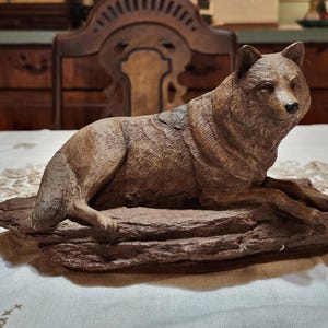 May include: A detailed wooden sculpture of a wolf in a resting pose. The wolf is brown with textured fur, lying on a dark brown, textured base. The sculpture is a decorative item, likely for home decor.