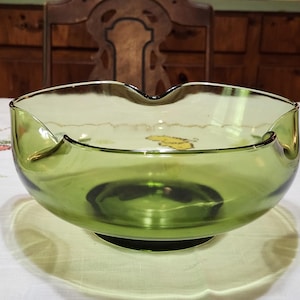 May include: A green glass bowl with a scalloped rim. The bowl has a wide, shallow design and sits on a small, round base. The glass has a slight tint, and the bowl is likely vintage or antique.