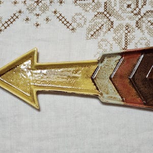 May include: A ceramic arrow-shaped serving dish with a textured surface. The dish features a gradient of colors, starting with yellow and transitioning to white, orange, and brown. The arrow is on a white surface.