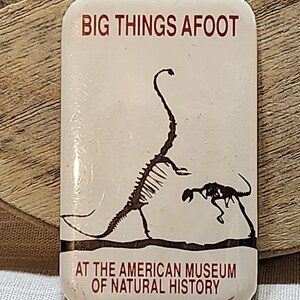May include: A rectangular, off-white pin with the text "BIG THINGS AFOOT" and "AT THE AMERICAN MUSEUM OF NATURAL HISTORY" in red. The pin features a black illustration of two dinosaur skeletons.