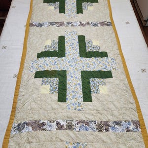 May include: A quilted table runner with a cross design. The crosses are made of green and floral fabric, with a beige background. The edges are trimmed with a gold border. The runner is approximately 60 inches long.