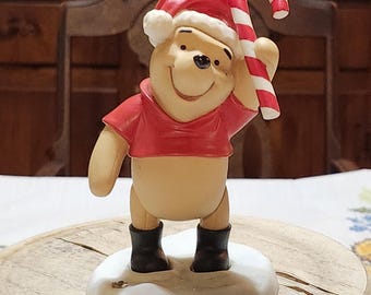 Peut inclure: Figurine de Winnie l'Ourson portant un bonnet de Père Noël et une chemise rouge, tenant un sucre d'orge. La figurine est sur une base blanche avec des bottes noires. Décoration de Noël.