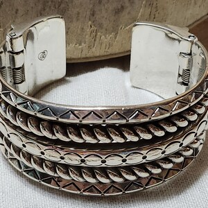 K&ouml;nnte beinhalten: Ein silbernes Armreif-Armband mit einem Klappverschluss. Das Armband hat drei Reihen detaillierter Muster, darunter Dreiecke, gedrehtes Seil und blattartige Formen. Das Metall hat eine polierte Oberfl&auml;che.