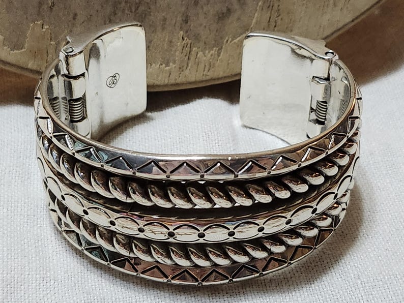 K&ouml;nnte beinhalten: Ein silbernes Armreif-Armband mit einem Klappverschluss. Das Armband hat drei Reihen detaillierter Muster, darunter Dreiecke, gedrehtes Seil und blattartige Formen. Das Metall hat eine polierte Oberfl&auml;che.
