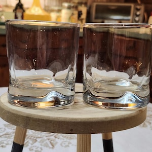 May include: Two clear, lowball glasses with a curved base sit side-by-side on a small, round wooden stool. The glasses are empty, and the background is a blurred kitchen scene with wooden cabinets and a countertop.