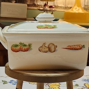 May include: Cream-colored rectangular ceramic casserole dish with a lid. The dish features painted illustrations of tomatoes, garlic, and a carrot. The lid has a small, round handle and a decorative border.