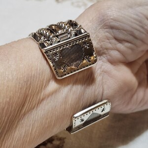 K&ouml;nnte beinhalten: Silberfarbenes Armreif-Armband mit aufwendigen Details. Der Hauptteil zeigt ein Herzdesign mit vertikalen Linien und sternf&ouml;rmigen Akzenten. Die Oberseite hat ein strukturiertes Design. Das Armband ist offen und verstellbar.