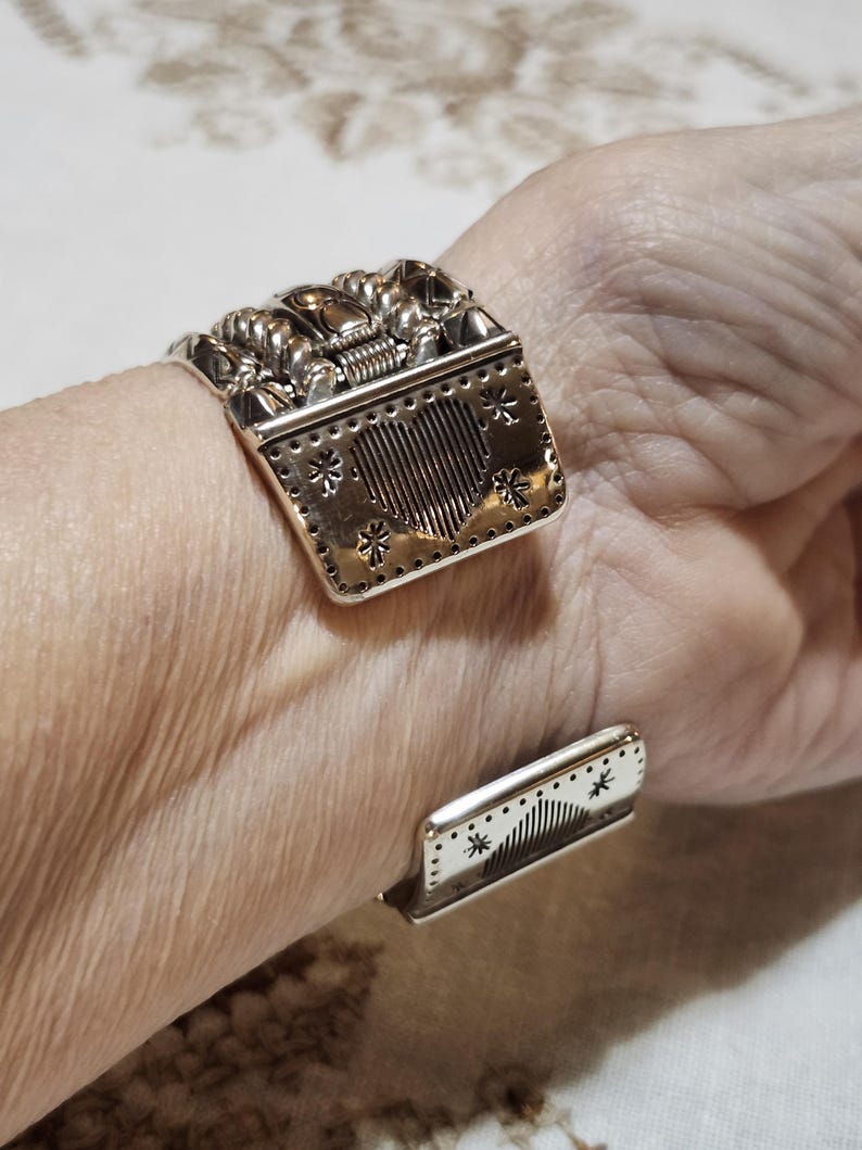 K&ouml;nnte beinhalten: Silberfarbenes Armreif-Armband mit aufwendigen Details. Der Hauptteil zeigt ein Herzdesign mit vertikalen Linien und sternf&ouml;rmigen Akzenten. Die Oberseite hat ein strukturiertes Design. Das Armband ist offen und verstellbar.